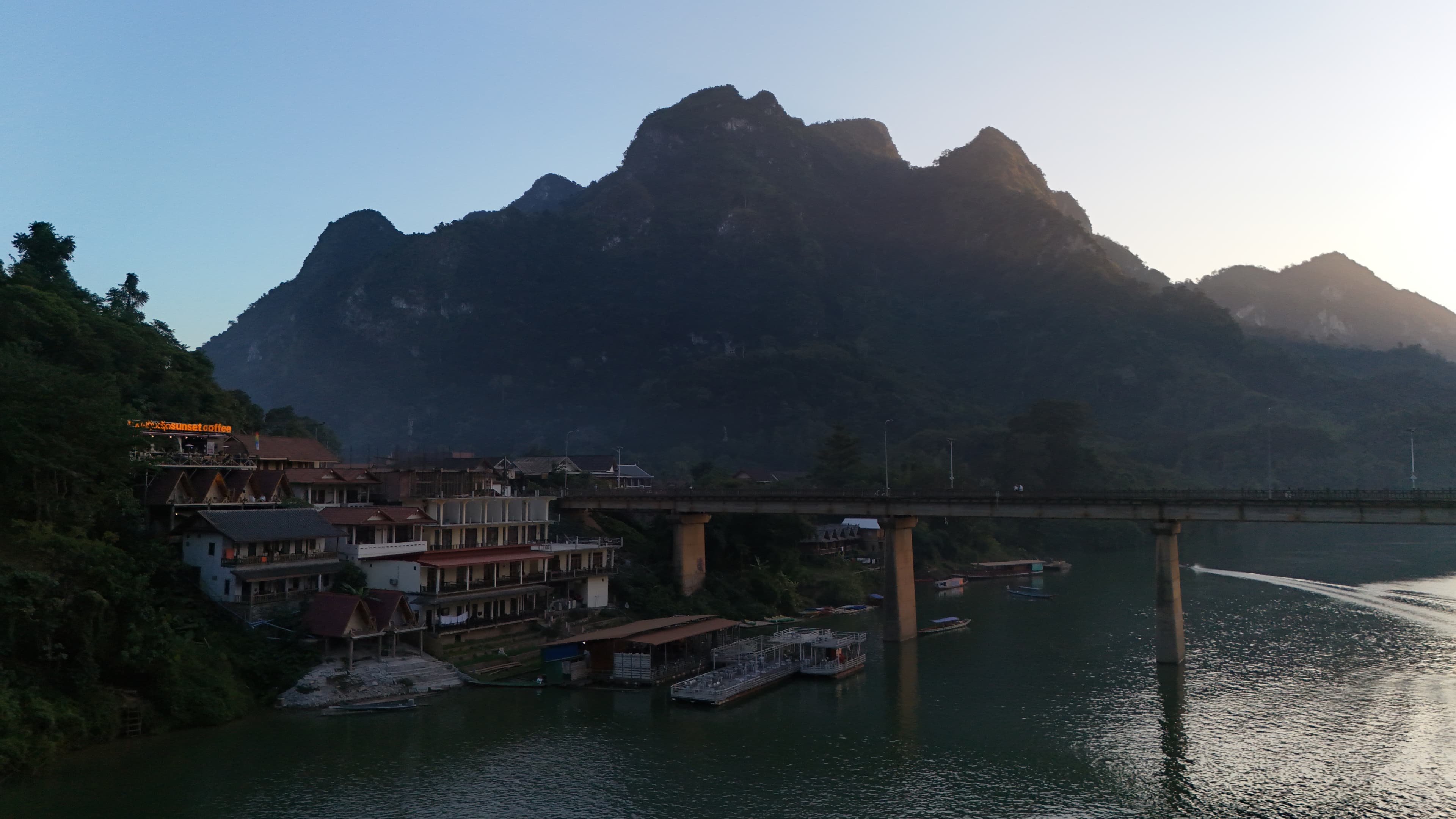 Drone view of a limestone karst peak rising behind Nong Khiaw, the Nam Ou river curving past the concrete bridge in late-afternoon light